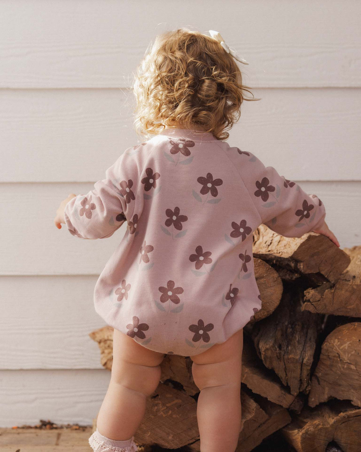 A toddler wearing the Rylee + Cru Daisy Crew Neck Romper in a soft pink hue with an all-over dark floral print.