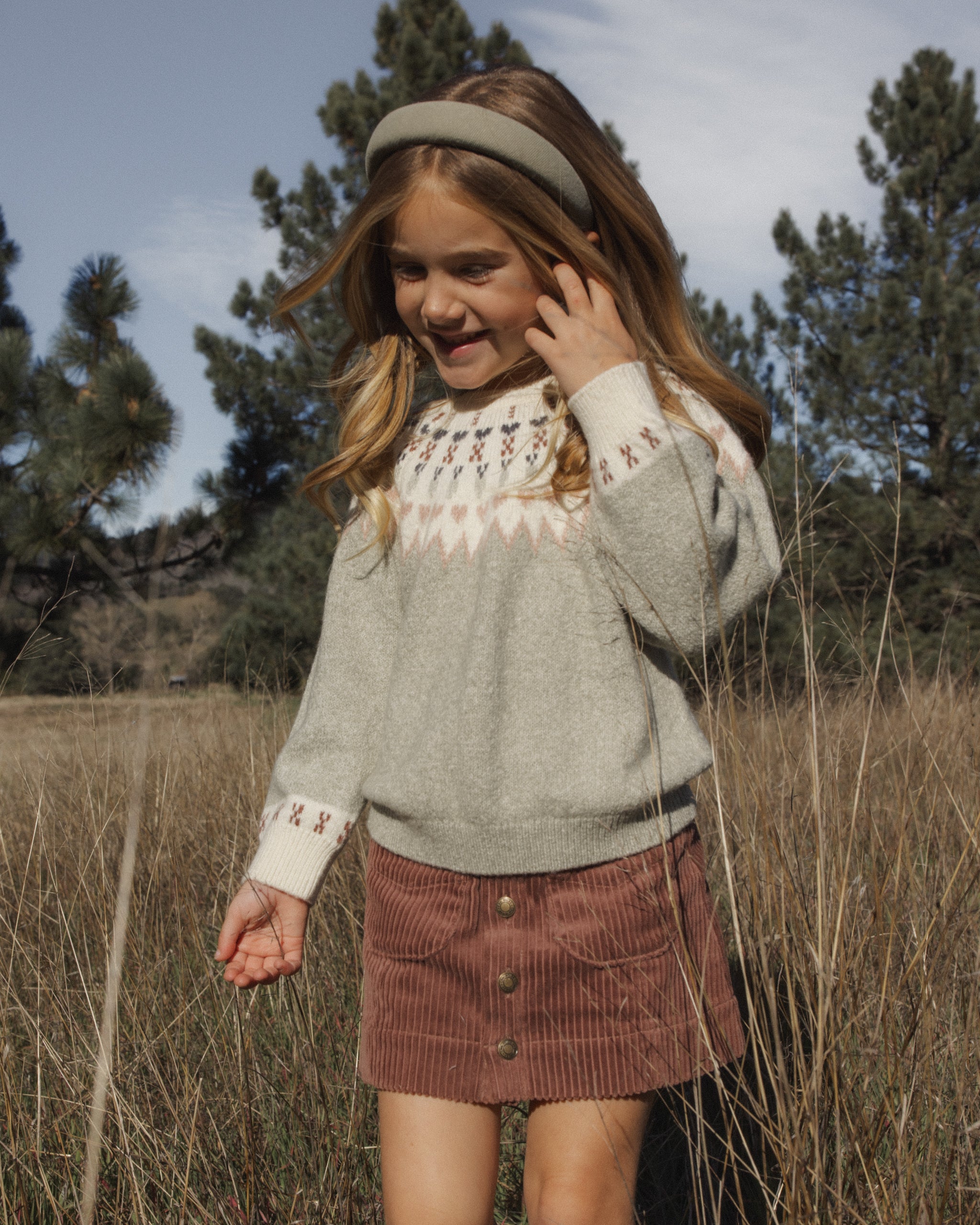 A young girl wearing a light gray knit sweater with a cream and red geometric pattern around the collar and cuffs, paired with a brown corduroy skirt, standing in a field of dry grass with trees in the background.