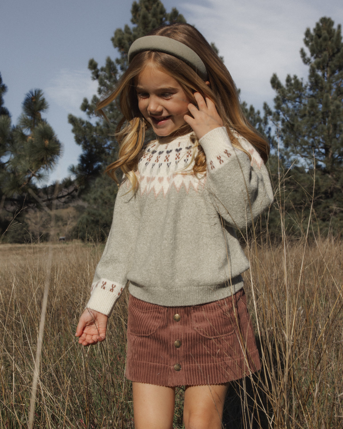 A young girl wearing a light gray knit sweater with a cream and red geometric pattern around the collar and cuffs, paired with a brown corduroy skirt, standing in a field of dry grass with trees in the background.