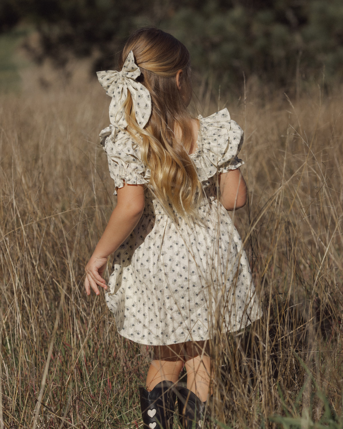 A young girl with long blonde hair is seen from behind, wearing a cream-colored dress with a small floral pattern and a large matching hair bow, standing in a field of dry grass.