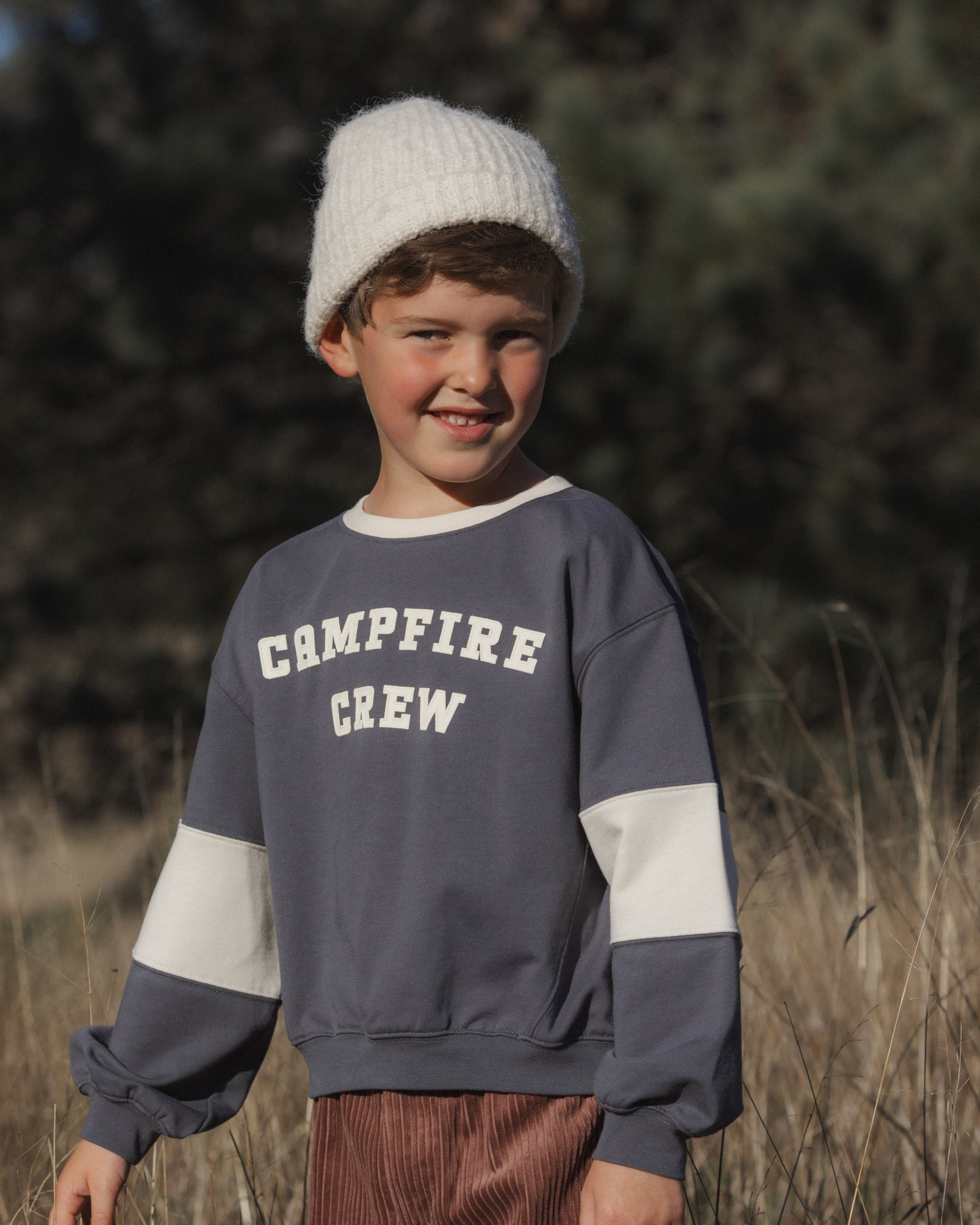 A young boy wearing a gray and white color-block crewneck sweatshirt with 'CAMPFIRE CREW' printed on the chest, paired with a white knit beanie and brown corduroy pants, standing outdoors in a grassy field.
