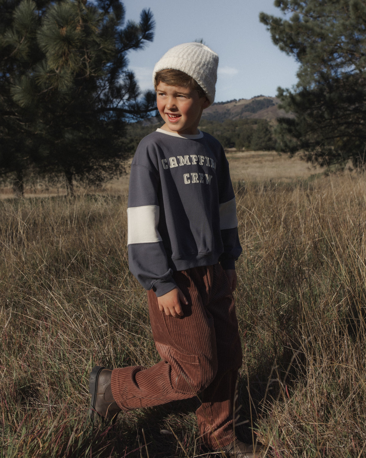 A young boy wearing a navy and white striped long-sleeve sweatshirt with 'CRIMSON CROW' printed on the chest, brown corduroy pants, and a white knit beanie, standing in a grassy field with trees in the background.