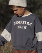 A child wearing a slate blue and white color-block sweatshirt with 'CAMPFIRE CREW' printed in white varsity lettering on the chest, paired with a white knit beanie.