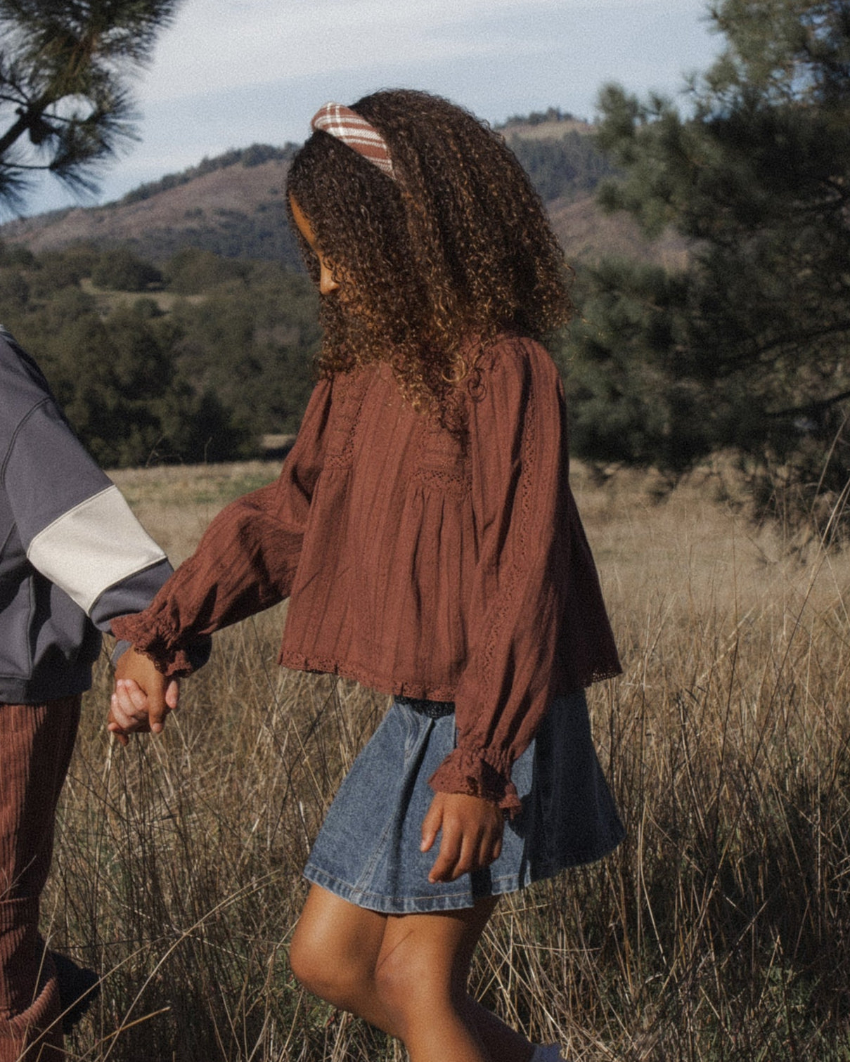 A child wearing a rust-brown long-sleeve textured blouse and a denim skirt walking through a dry grass field outdoors, holding hands with another person partially visible on the left.