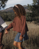 A child wearing a rust-brown long-sleeve textured blouse and a denim skirt walking through a dry grass field outdoors, holding hands with another person partially visible on the left.
