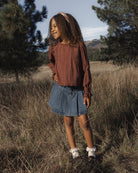 A young girl standing in an outdoor grassy field wearing a rust-brown long-sleeve top, a blue denim skirt, and white sneakers.