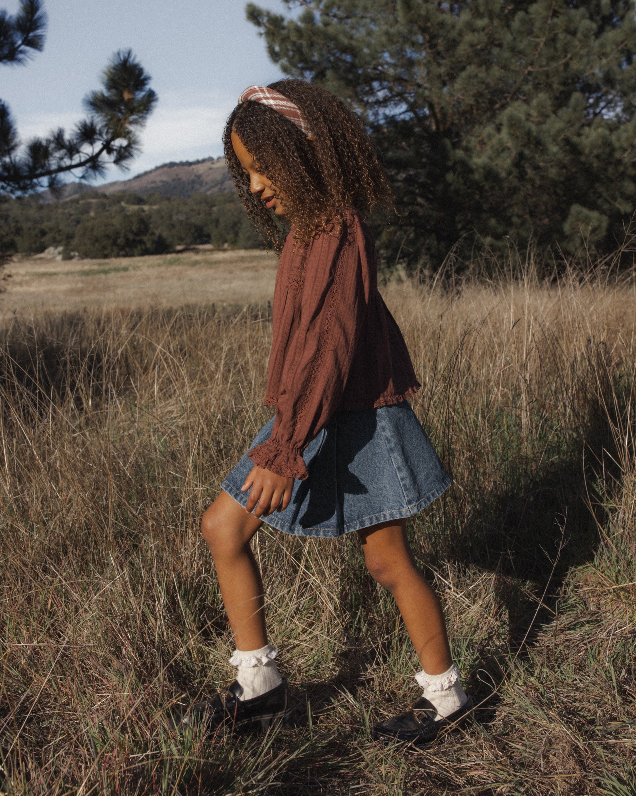 A young girl walking through a dry grass field outdoors, wearing a brown long-sleeve top, a denim skirt, white socks, and black shoes.