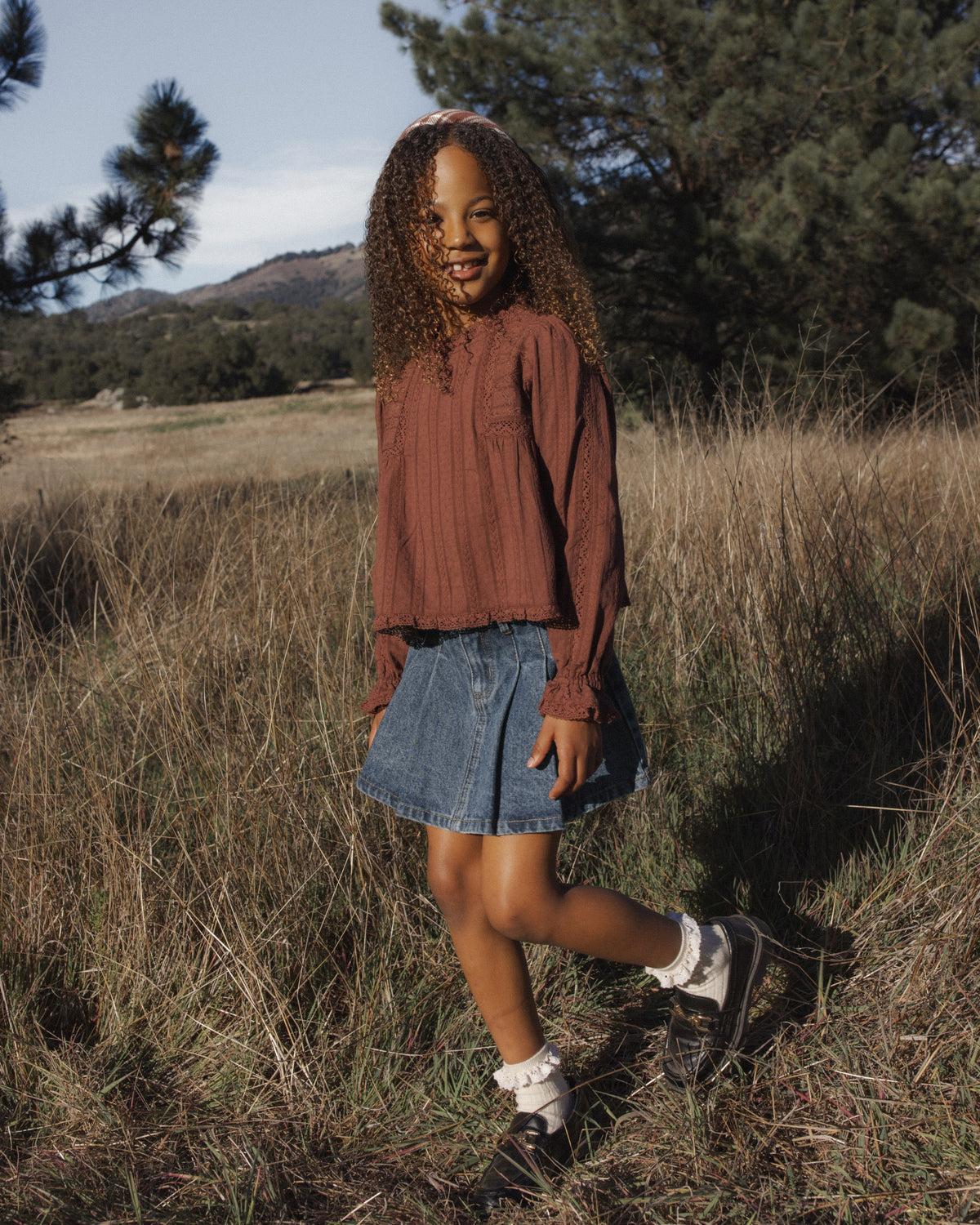 A woman standing outdoors in a grassy field wearing a rust-brown long-sleeve top, a denim mini skirt, white socks, and black sneakers.