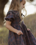 A young girl with long blonde hair wearing a dark blue and red plaid dress, standing outdoors in a softly blurred natural setting.