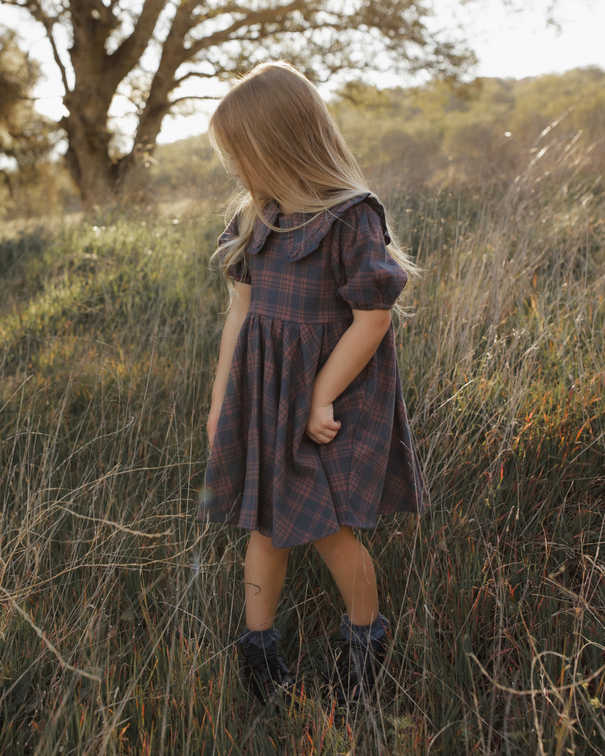A young girl with long blonde hair stands in a grassy outdoor field wearing a dark plaid short-sleeve dress with a large ruffled collar and dark boots.