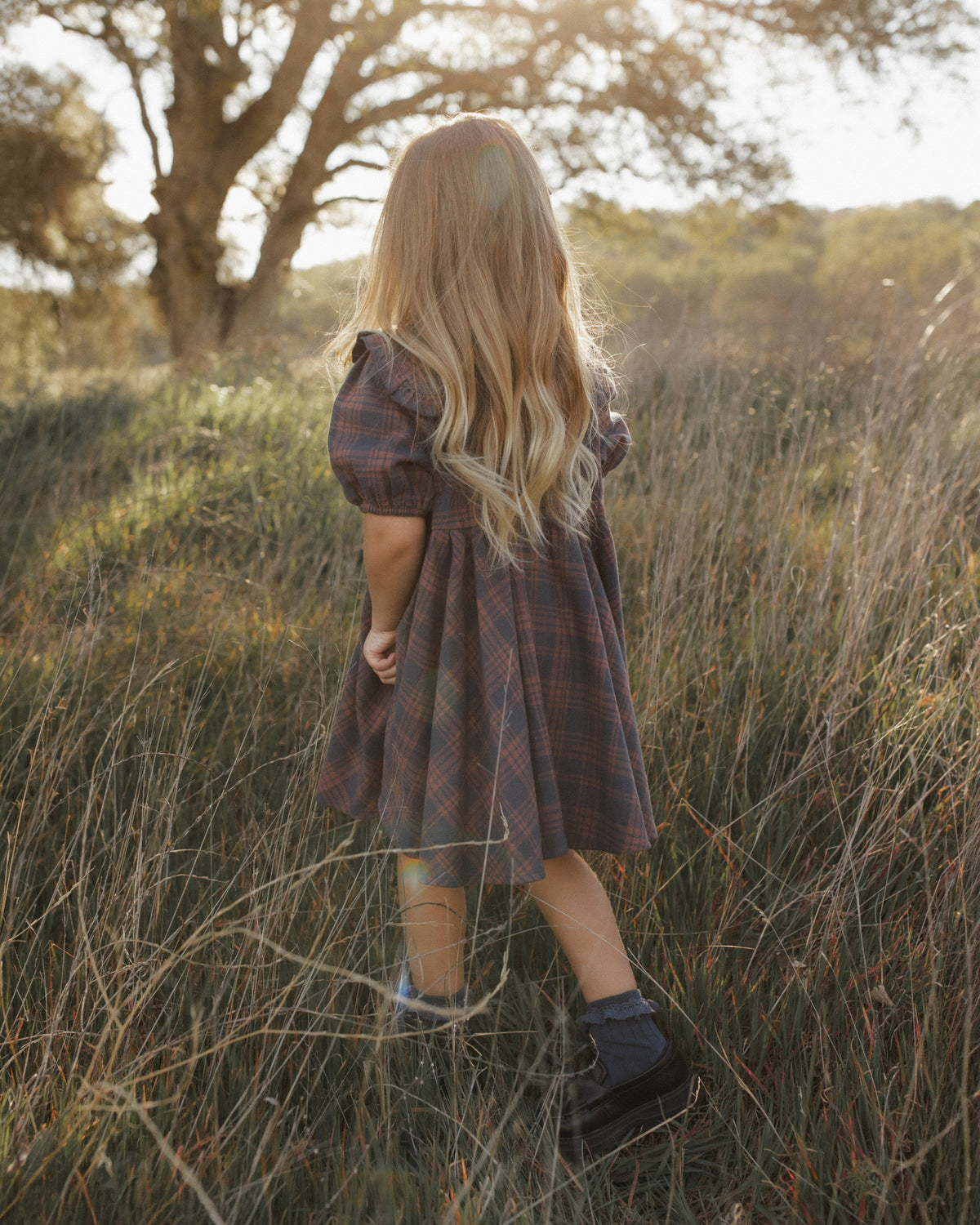 A young girl with long blonde hair stands in a grassy outdoor field, viewed from behind. She is wearing a dark plaid dress with puff sleeves and dark socks with boots.