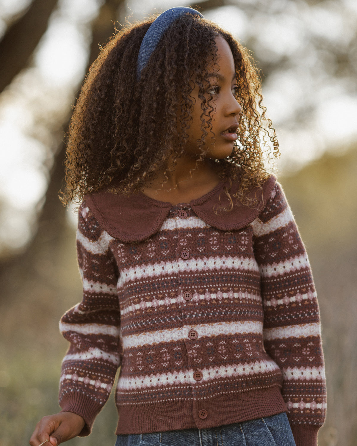 A young girl wearing a brown and cream patterned knit cardigan sweater with a Peter Pan collar, photographed outdoors with a blurred natural background.