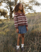 A young girl stands outdoors in a grassy field wearing a brown and white striped knit sweater, a denim skirt, white socks, and dark boots.