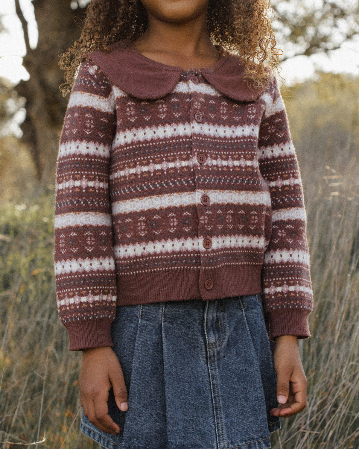 A child wearing a brown and cream patterned knit cardigan with a large brown Peter Pan collar, paired with a blue denim skirt, standing outdoors in a field.