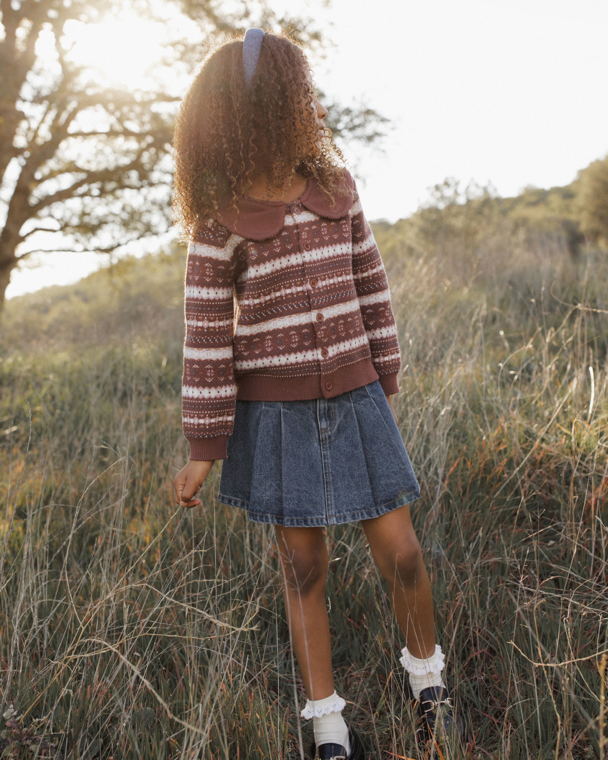 A young girl standing outdoors in a grassy field, viewed from behind, wearing a brown and cream patterned cardigan with a large collar, a blue denim skirt, white socks, and black shoes.