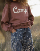A child wearing a brownish-rose crewneck sweatshirt with 'Camp' written in white script across the chest, paired with blue patterned pants, standing outdoors in a field.