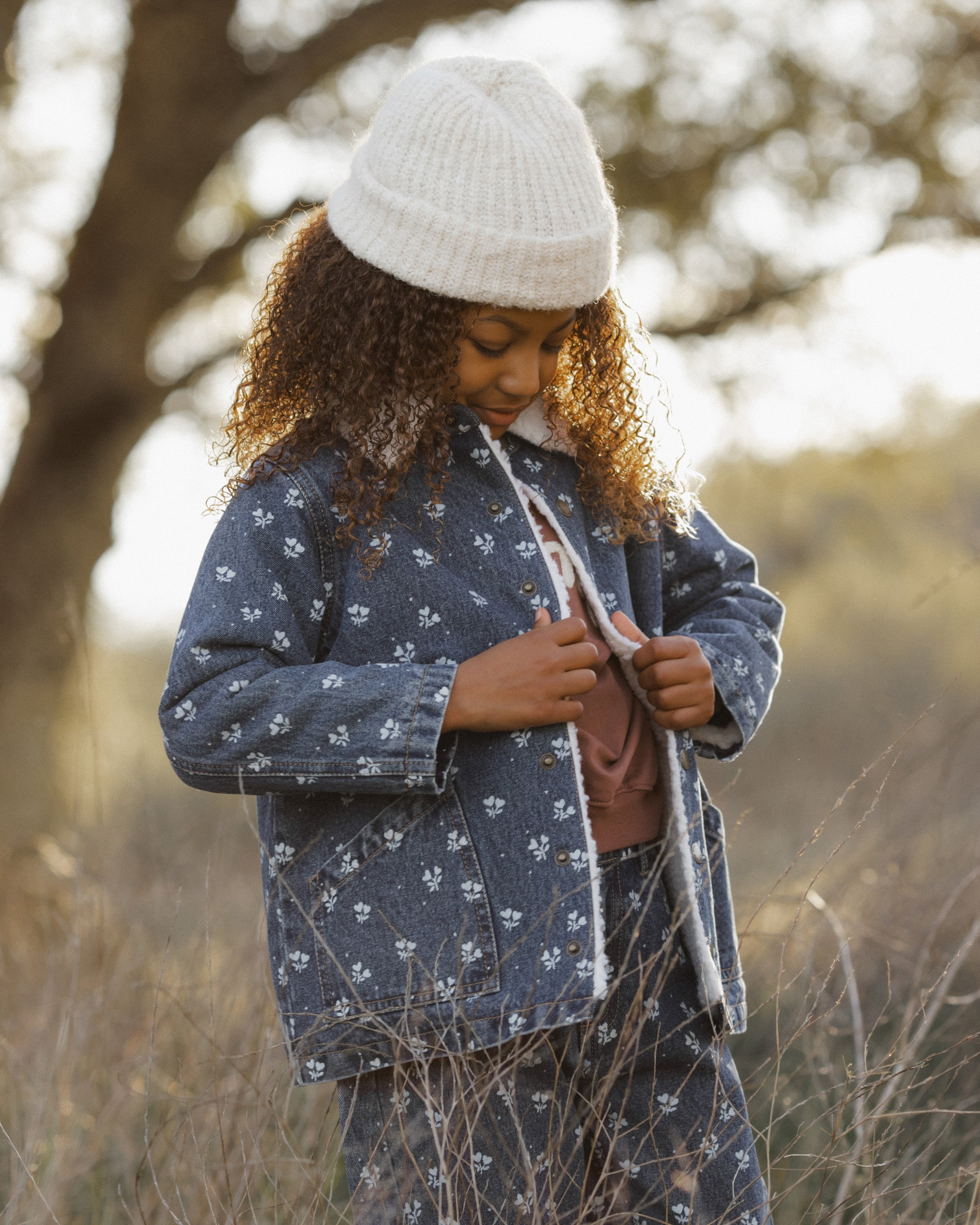 A child wearing a matching navy blue pajama set with a white floral print, featuring a long-sleeve top and pants, styled with a white knit beanie in an outdoor field.