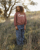 A person standing outdoors in a grassy field wearing a dusty rose-brown sweatshirt with 'Camp' printed in white across the chest, blue patterned pants, and a white cap.