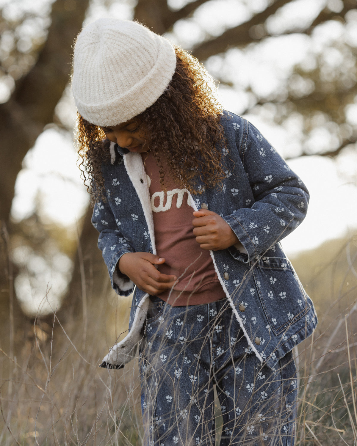 A child wearing a matching blue patterned pajama set with a white knit beanie, standing outdoors in a grassy field.