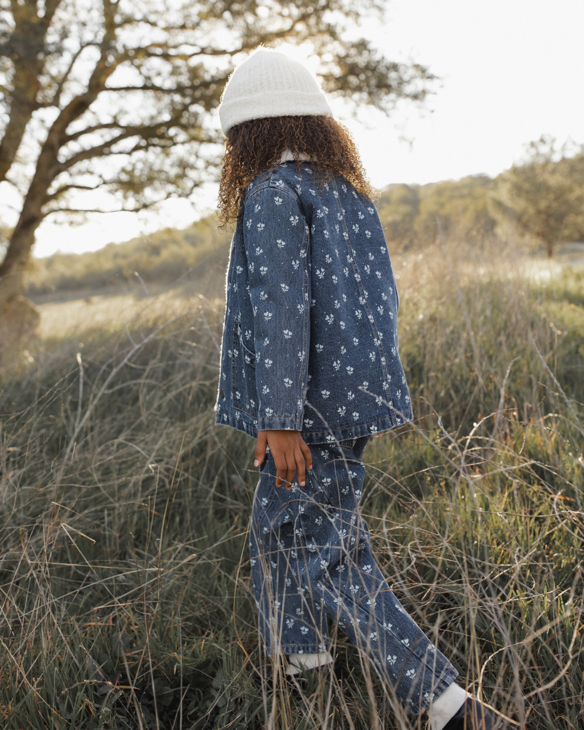 A child wearing a matching blue denim-like pajama set with a small white floral print and a white knit beanie, standing in tall dry grass outdoors near trees.