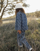 A child wearing a matching blue denim-like pajama set with a small white floral print and a white knit beanie, standing in tall dry grass outdoors near trees.
