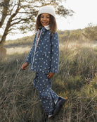 A young child wearing a blue patterned long-sleeve top and matching pants with a white beanie, standing outdoors in a grassy field.
