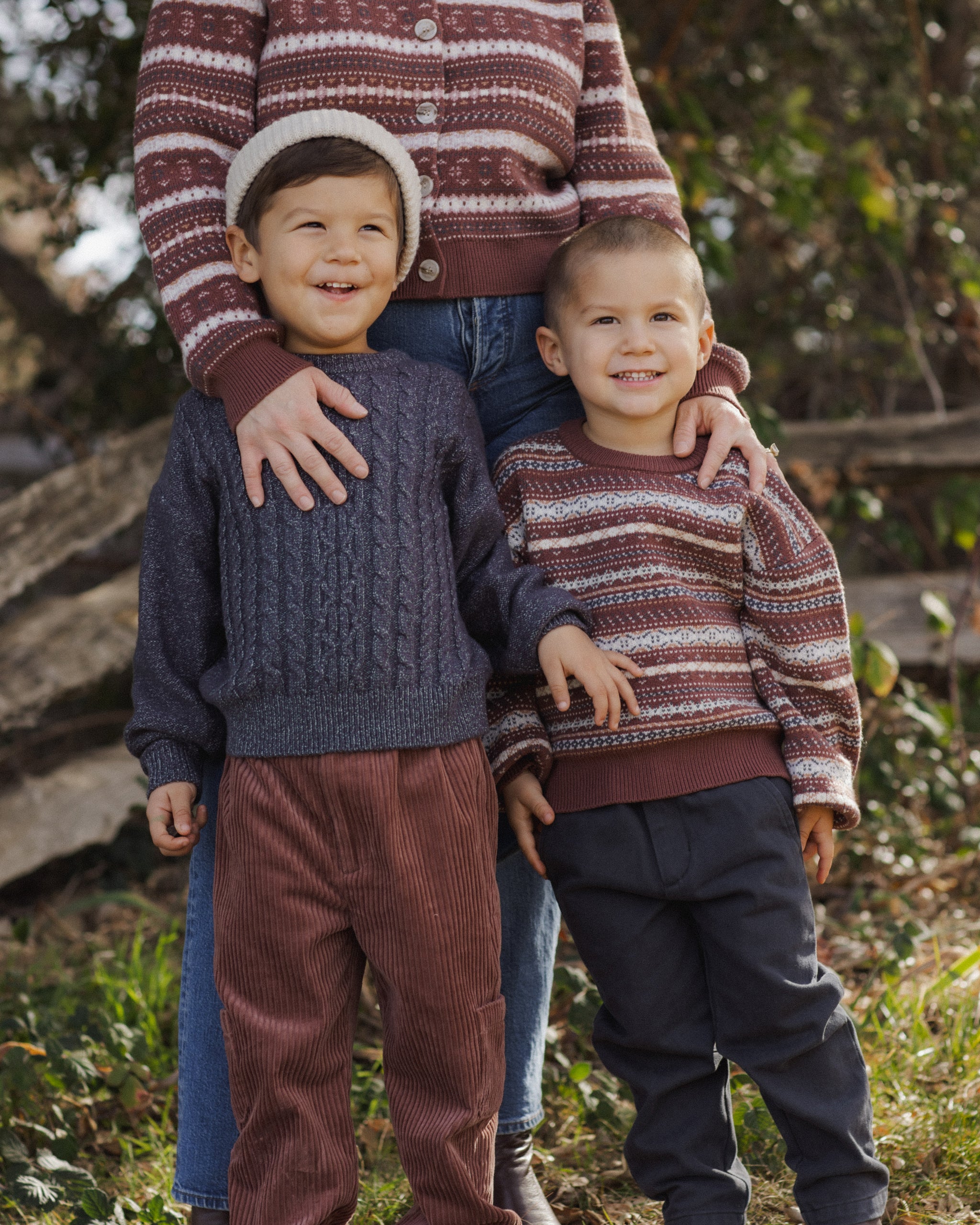 Two children wearing matching striped sweaters standing outdoors with a person partially visible.