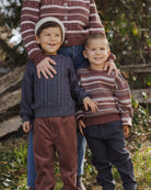Two children wearing matching striped sweaters standing outdoors with a person partially visible.