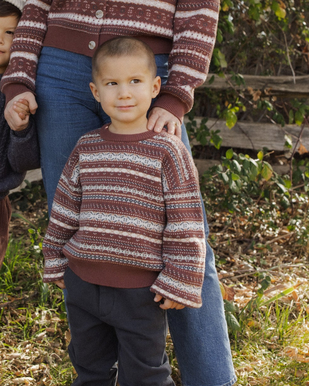 A woman and two young boys wearing matching knit sweaters with a rust brown and cream horizontal stripe pattern, standing outdoors in a wooded setting.