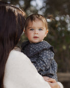 A toddler with blue eyes and a small ponytail wearing a dark gray patterned dress, held by an adult with long dark hair in a white sweater, set against a soft-focus outdoor background.