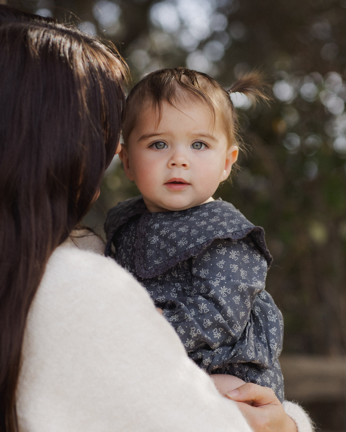 A toddler with blue eyes and a small ponytail wearing a dark gray patterned dress, held by an adult with long dark hair in a white sweater, set against a soft-focus outdoor background.