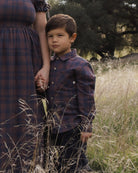 A young boy wearing a dark blue and brown plaid button-down shirt and dark pants stands in a grassy outdoor field next to an adult woman in a matching plaid dress.