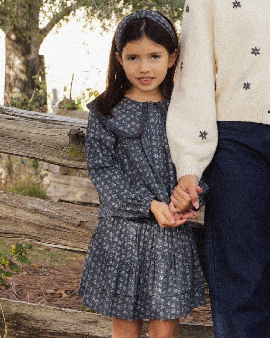 A young girl wearing a dark blue patterned long-sleeve dress with a large Peter Pan collar, standing outdoors next to an adult wearing a cream sweater and navy pants.