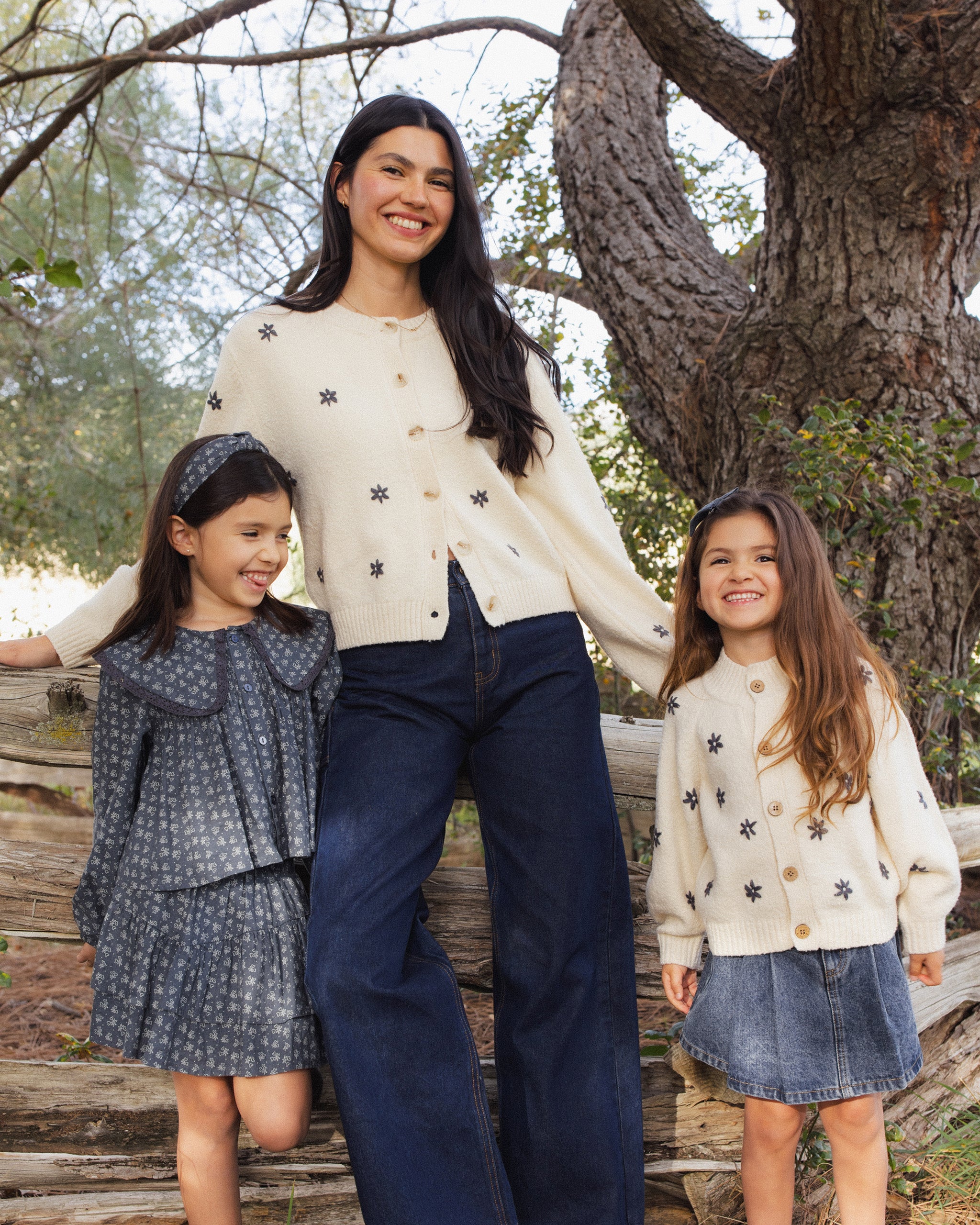 A woman and two young girls standing outdoors in front of a large tree, wearing coordinated knit clothing. The woman wears a cream knit top with small dark motifs and dark wide-leg pants, while the girls wear matching cream and gray outfits.