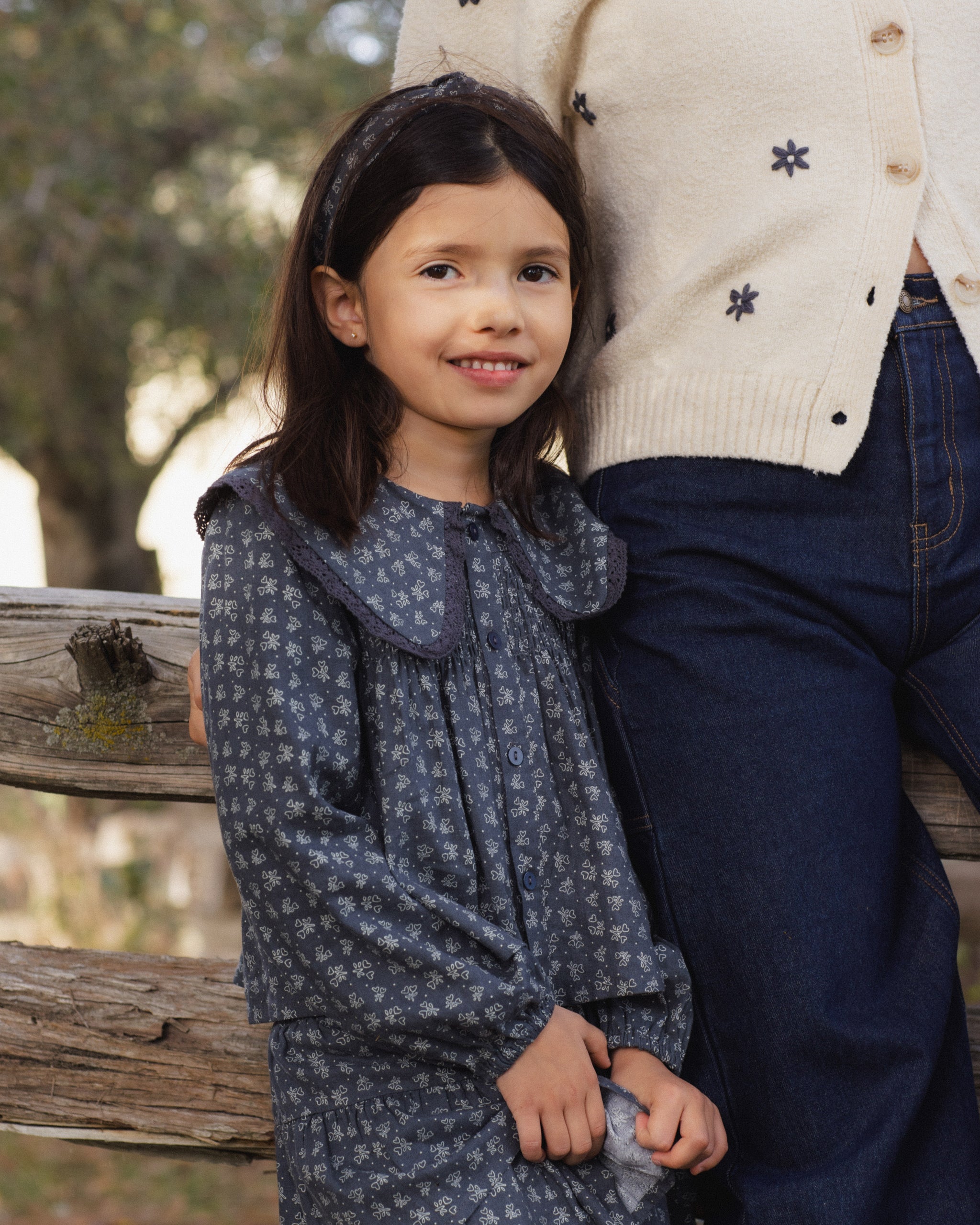 A young girl wearing a dark blue patterned long-sleeve dress stands next to an adult wearing a cream sweater and dark blue jeans, outdoors against a rustic wooden fence.