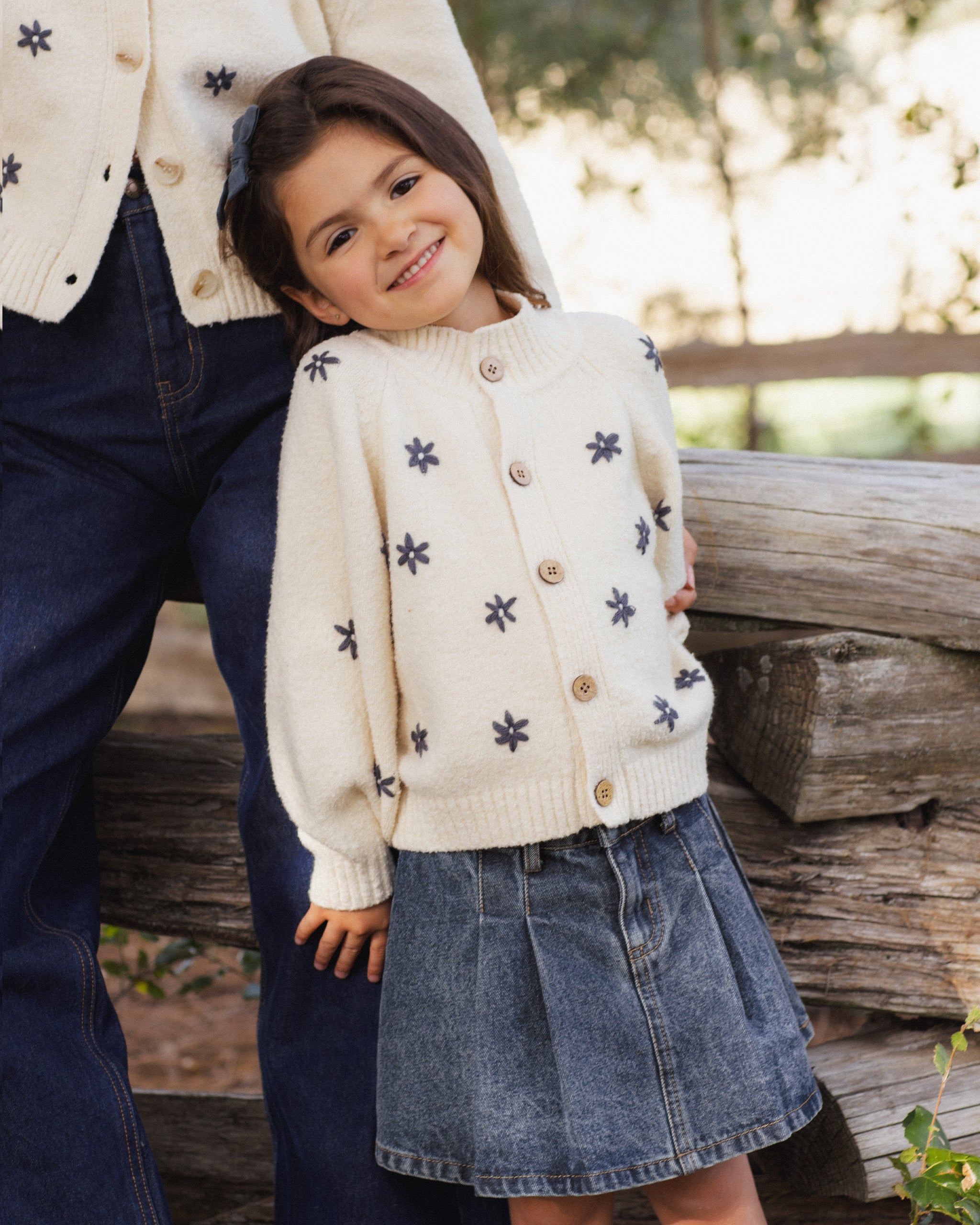 A young girl wearing a cream knit cardigan with dark blue star motifs and wooden buttons, styled with a denim skirt, standing next to an adult wearing a matching cream cardigan and dark jeans in an outdoor setting.