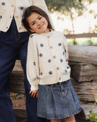 A young girl wearing a cream knit cardigan with dark blue star motifs and wooden buttons, styled with a denim skirt, standing next to an adult wearing a matching cream cardigan and dark jeans in an outdoor setting.