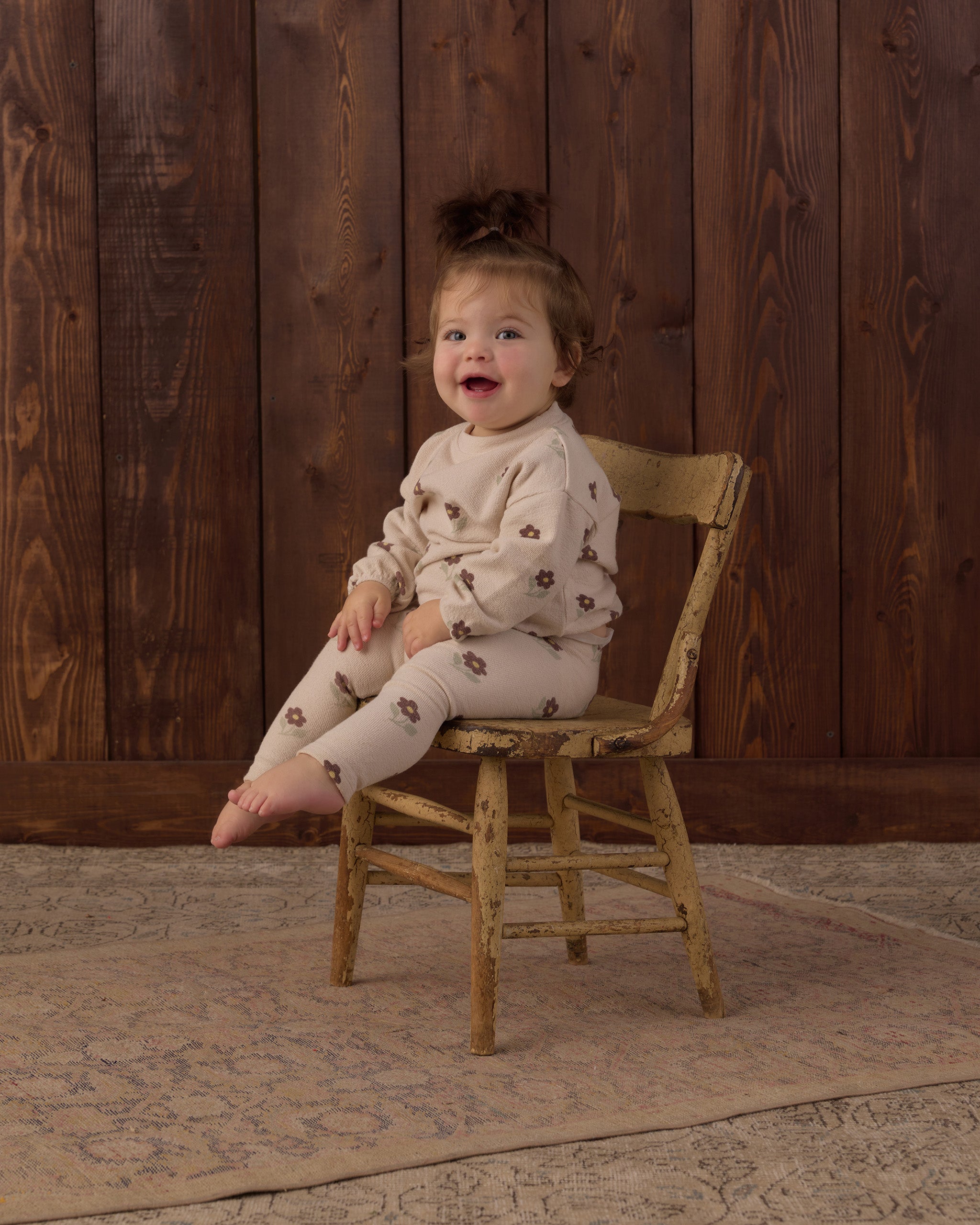 A toddler girl with a small hair bun sits on a rustic wooden chair, wearing a cream-colored long-sleeve pajama set with a brown floral pattern. The background consists of dark brown wooden wall panels and a patterned rug.