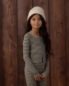 A young girl with long dark hair stands against a dark brown wooden plank wall, wearing a cream knit beanie and a matching long-sleeve striped one-piece outfit with thin black and beige horizontal lines.