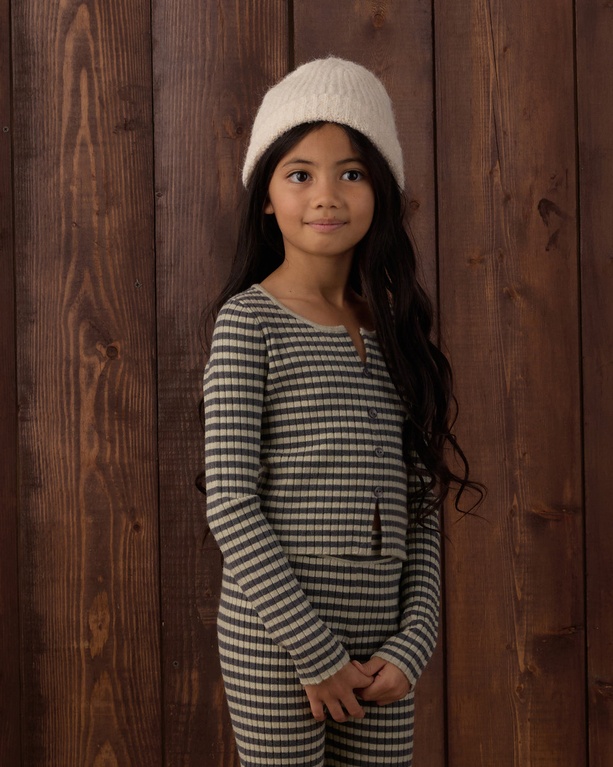 A young girl with long dark hair stands against a dark brown wooden plank wall, wearing a cream knit beanie and a matching long-sleeve striped one-piece outfit with thin black and beige horizontal lines.