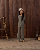 A young girl stands in front of a rustic dark wood plank wall wearing a matching black and white striped long-sleeve top and wide-leg pants set, paired with a white beanie and tan shoes.