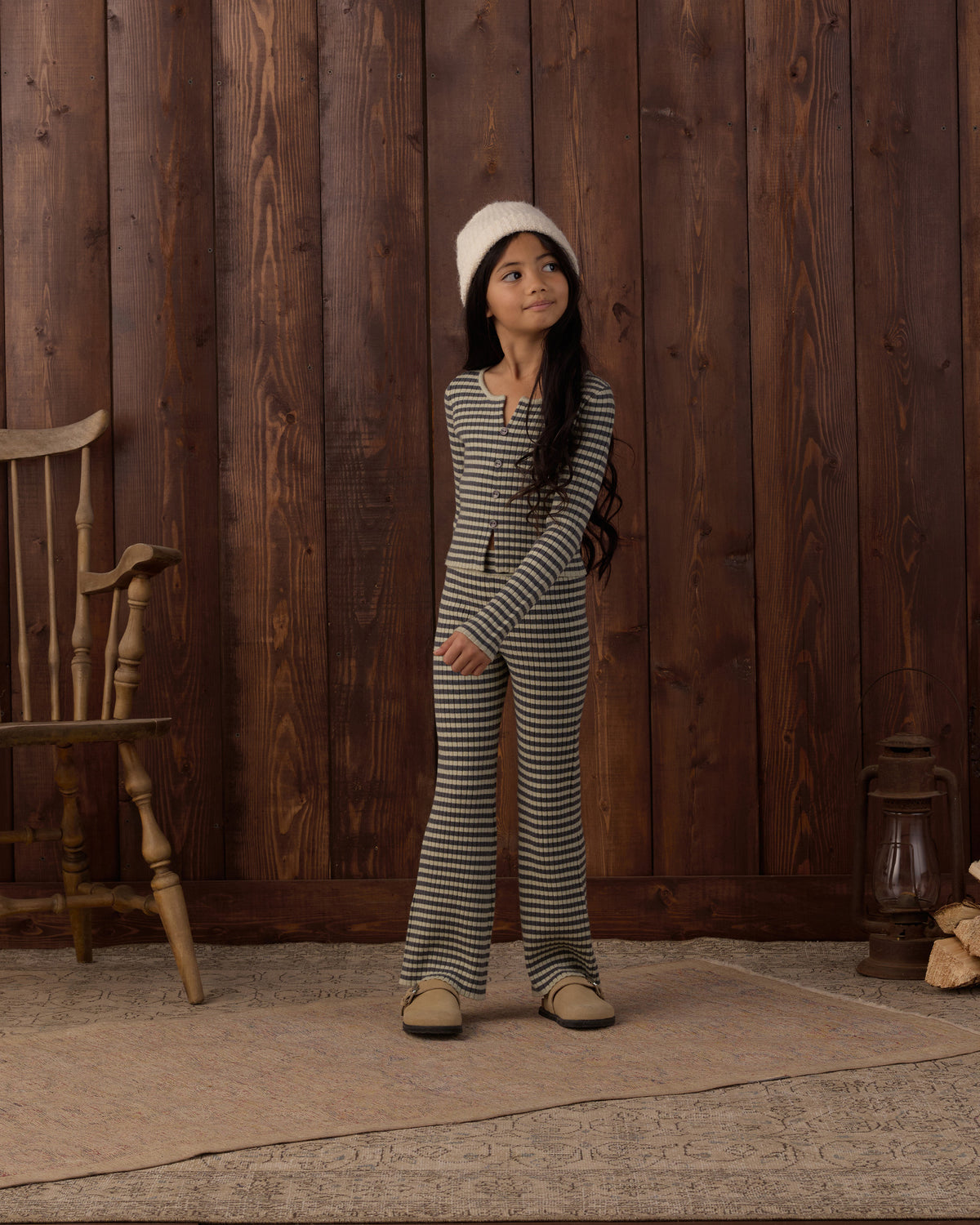 A young girl stands in front of a rustic dark wood plank wall wearing a matching black and white striped long-sleeve top and wide-leg pants set, paired with a white beanie and tan shoes.