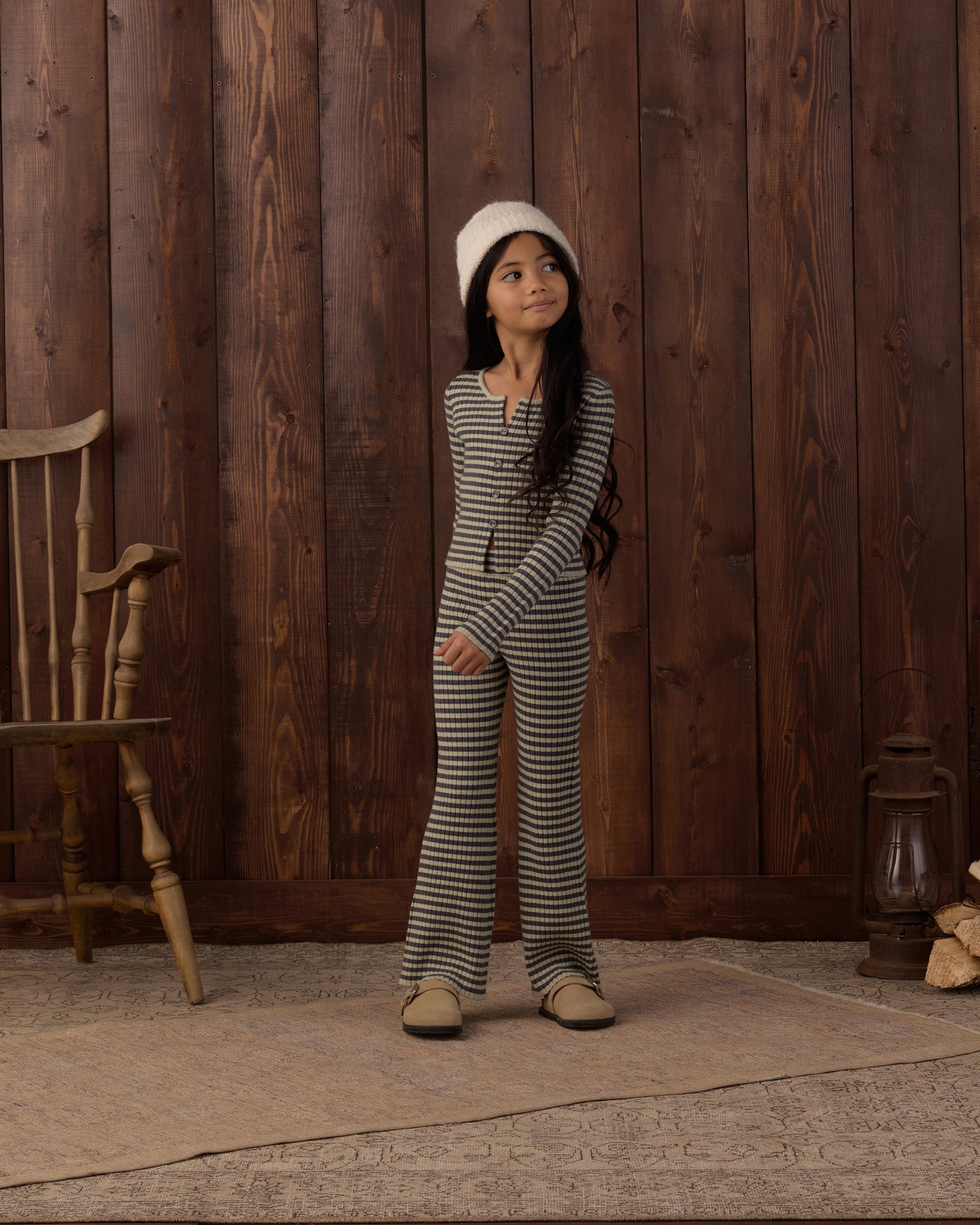A young girl stands in front of a rustic dark wood plank wall wearing a matching black and white striped long-sleeve top and wide-leg pants set, paired with a white beanie and tan shoes.