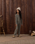 A young girl wearing a matching gray and white striped long-sleeve top and striped pants, styled with a cream knit beanie and beige shoes, standing in front of a rustic dark wood plank wall.