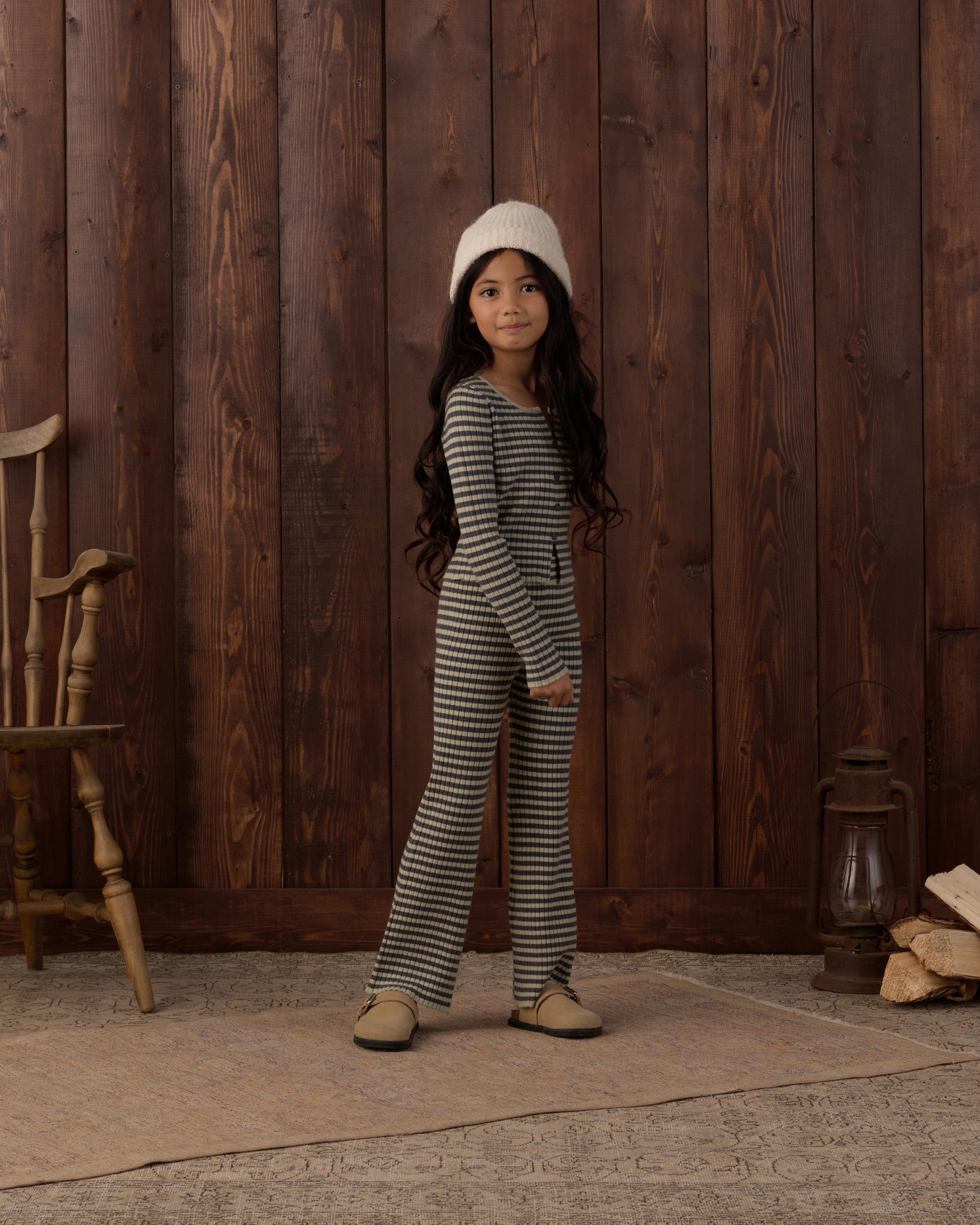 A young girl stands in front of a rustic dark wood plank wall wearing a matching long-sleeve striped one-piece outfit with a cream knit beanie and tan shoes.