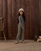 A young girl stands in front of a rustic dark wood plank wall wearing a matching long-sleeve striped one-piece outfit with a cream knit beanie and tan shoes.