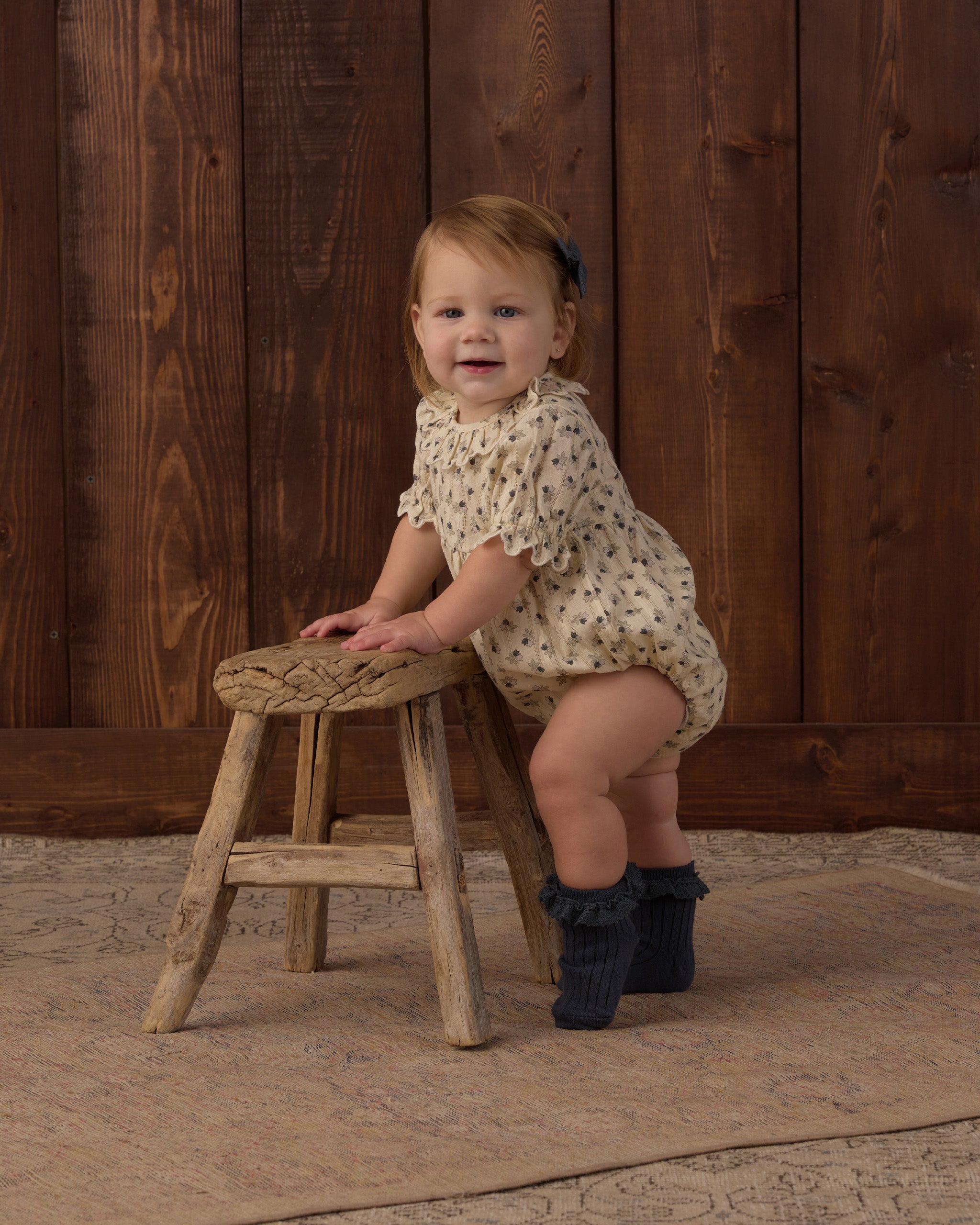A toddler girl wearing a cream short-sleeve romper with small dark polka dots and dark ruffled booties, sitting on a rustic wooden stool in front of a dark wood plank wall.