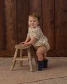 A toddler girl wearing a cream short-sleeve romper with small dark polka dots and dark ruffled booties, sitting on a rustic wooden stool in front of a dark wood plank wall.