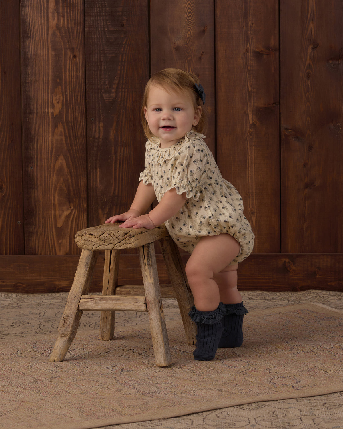 A toddler girl wearing a cream short-sleeve romper with small dark polka dots and dark ruffled booties, sitting on a rustic wooden stool in front of a dark wood plank wall.