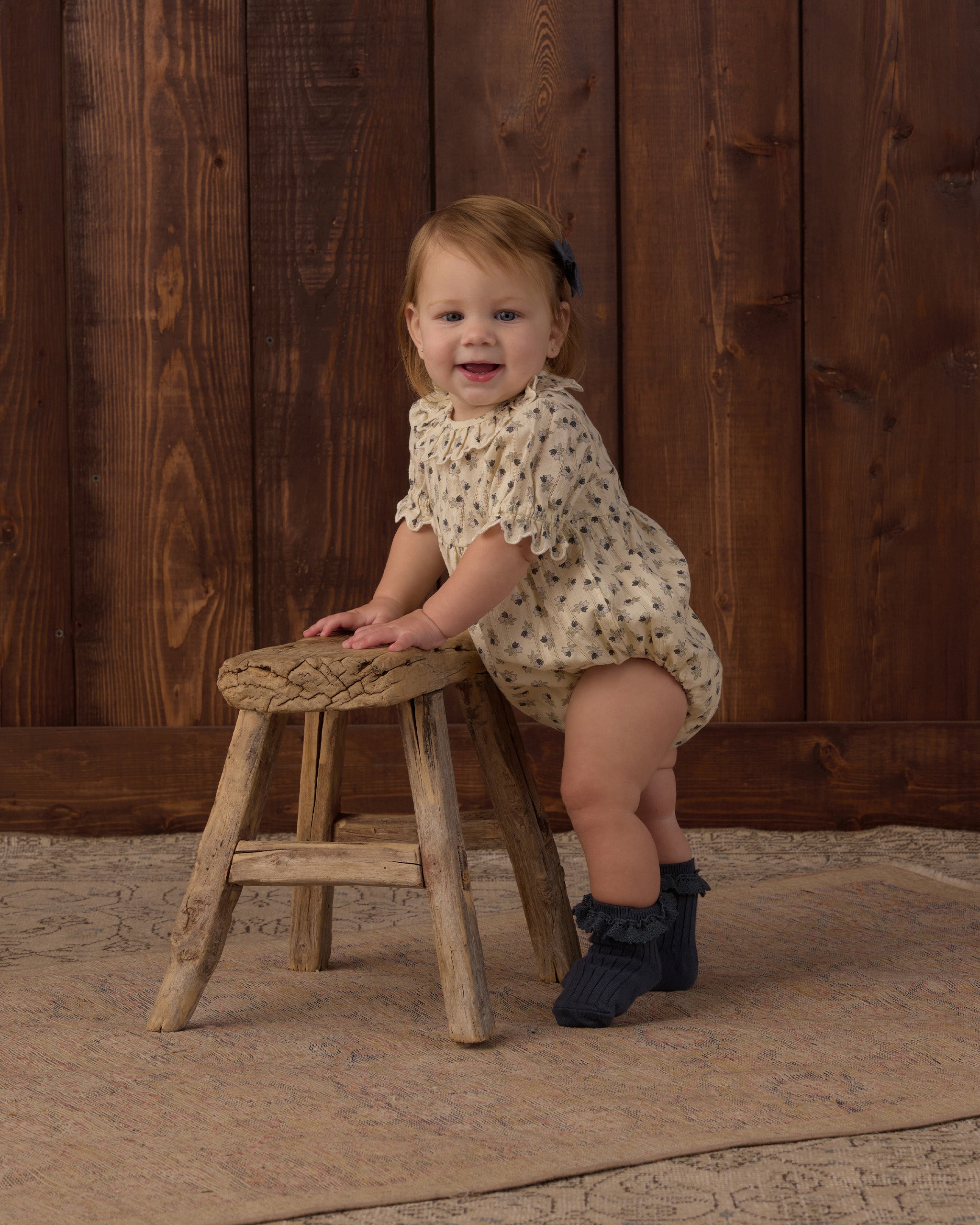 A toddler girl wearing a cream short-sleeve romper with a small floral print sits on a rustic wooden stool in front of a dark wood plank wall.